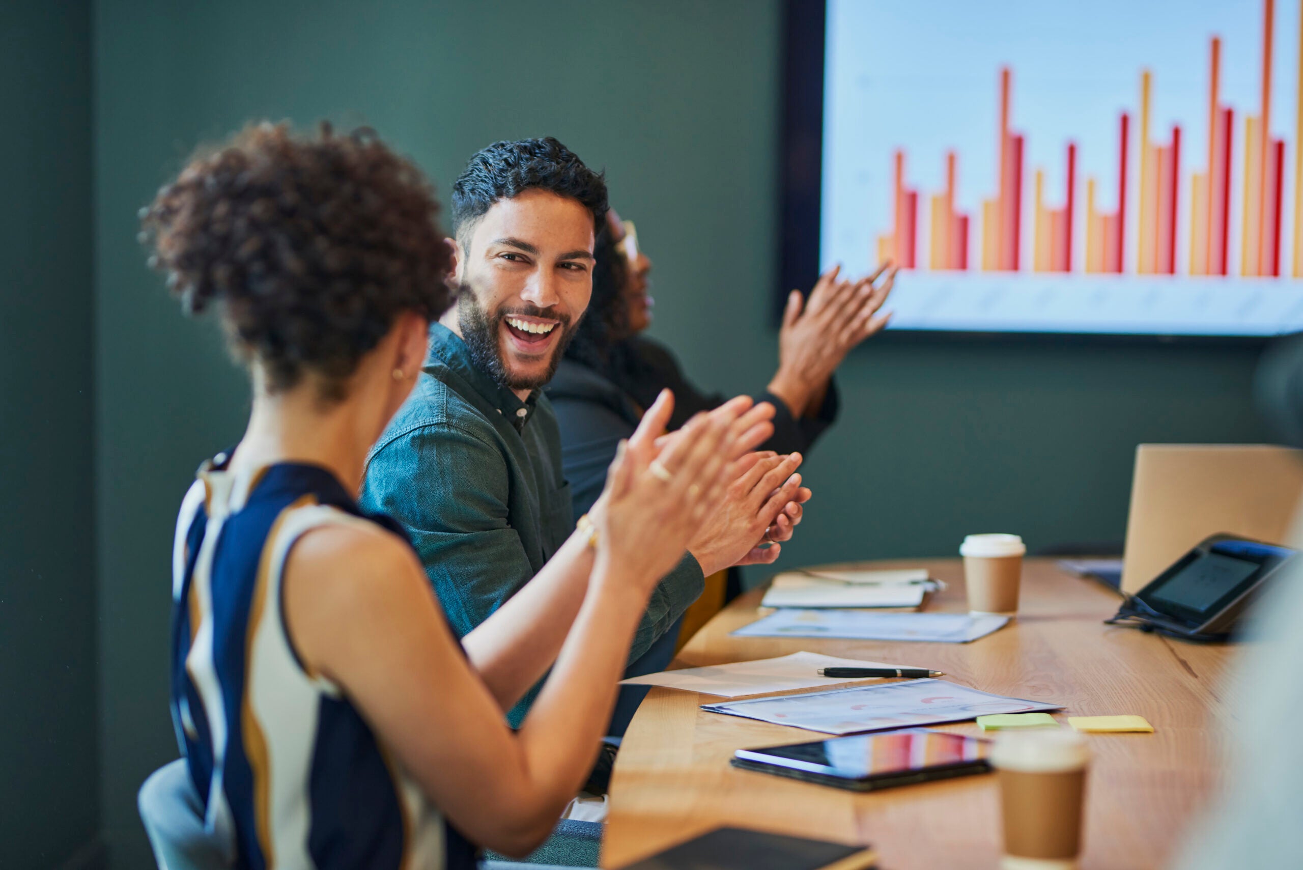 Group of business professionals clapping and smiling during a meeting in the boardroom. They are all sitting around a conference table and looking at a presentation on the screen. The people are all dressed in business casual attire and are engaged in the meeting.