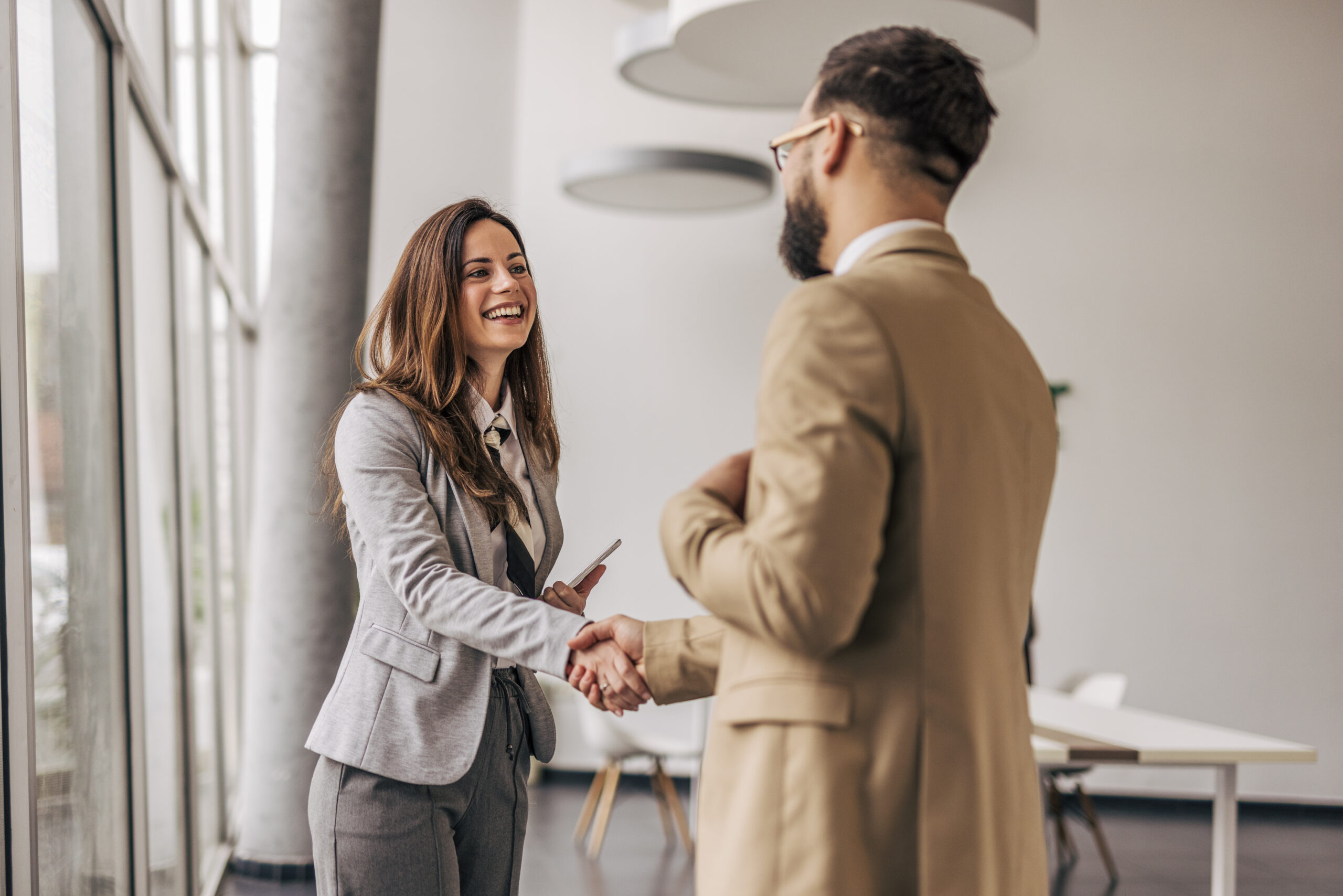 Two young business professionals shaking hands in a modern office, the woman smiling while holding a smartphone, celebrating a successful deal and partnership